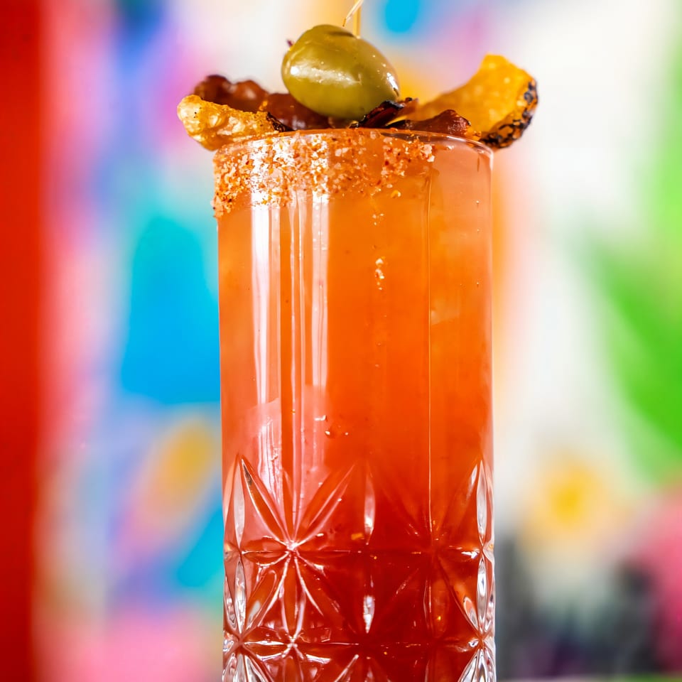 A tall, red cocktail in a textured glass, garnished with an olive, bacon, and a chili-lime rim.