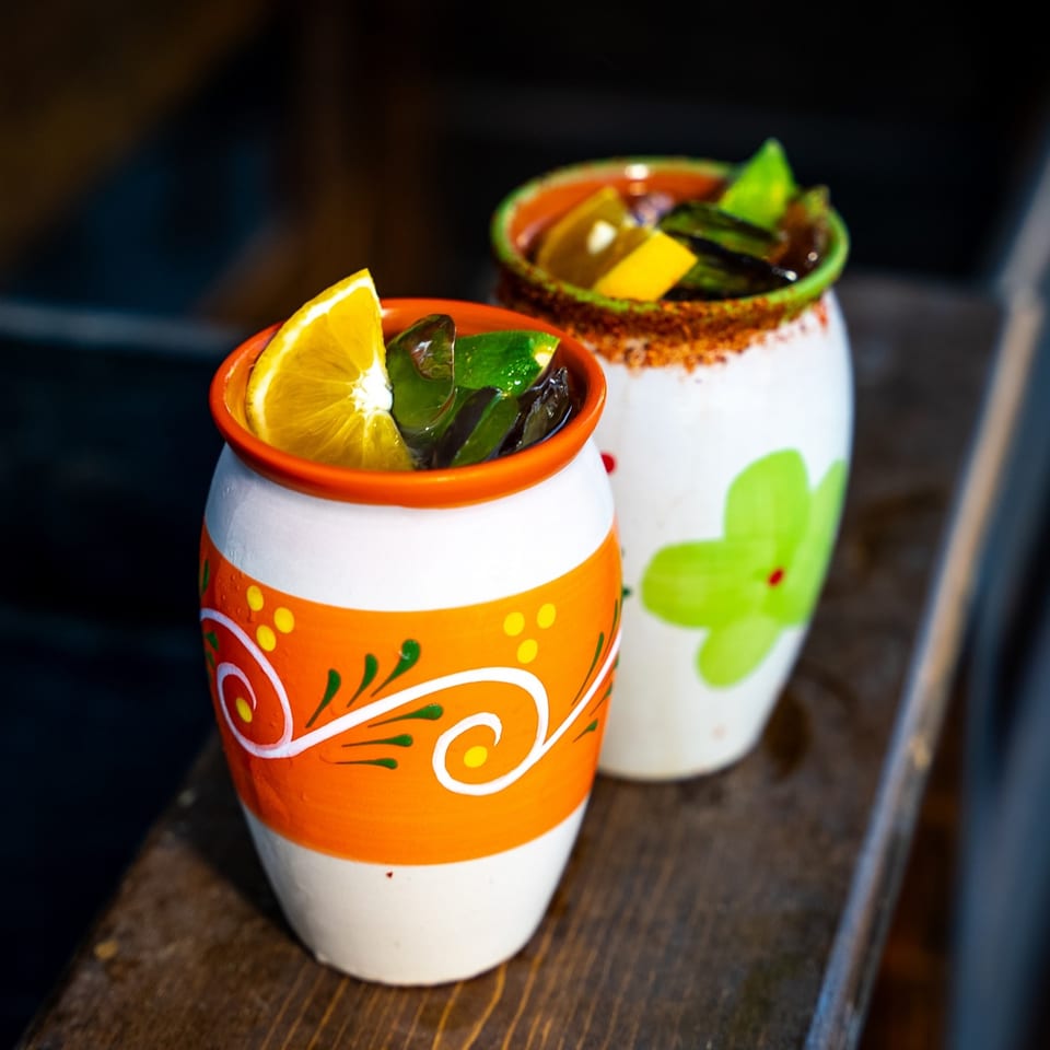Two colorful Mexican cocktails in ceramic mugs, garnished with citrus, on a wooden surface.