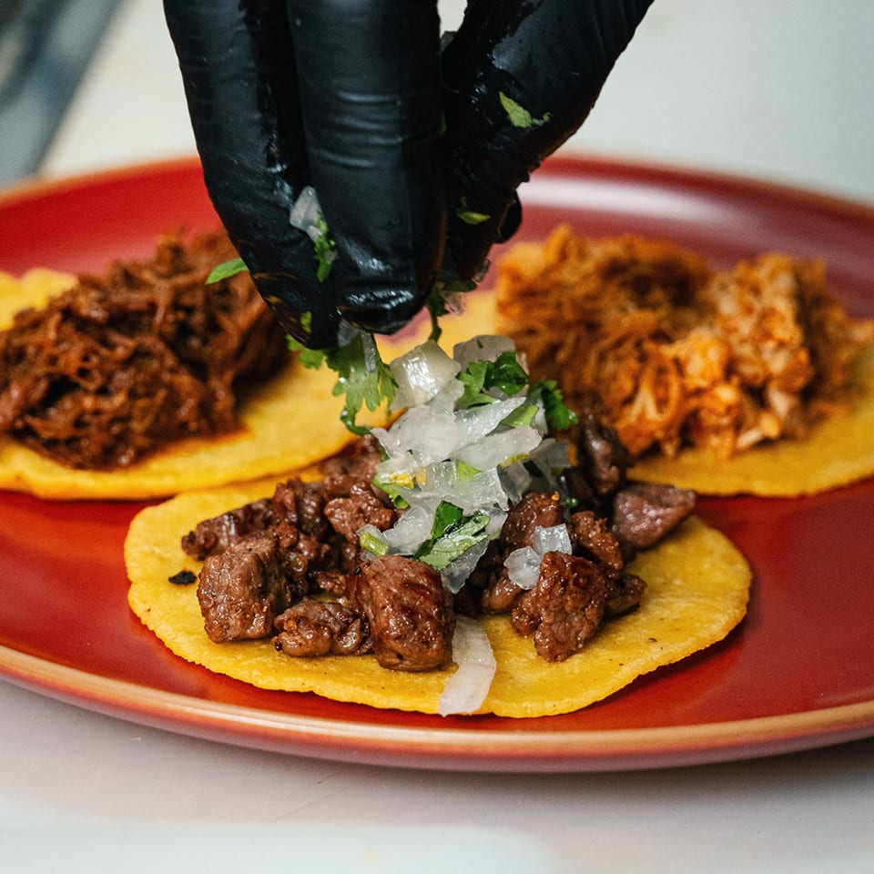 Gloved hands garnishing three Mi Barrio Kitchen & Cocktails tacos with onions and cilantro.