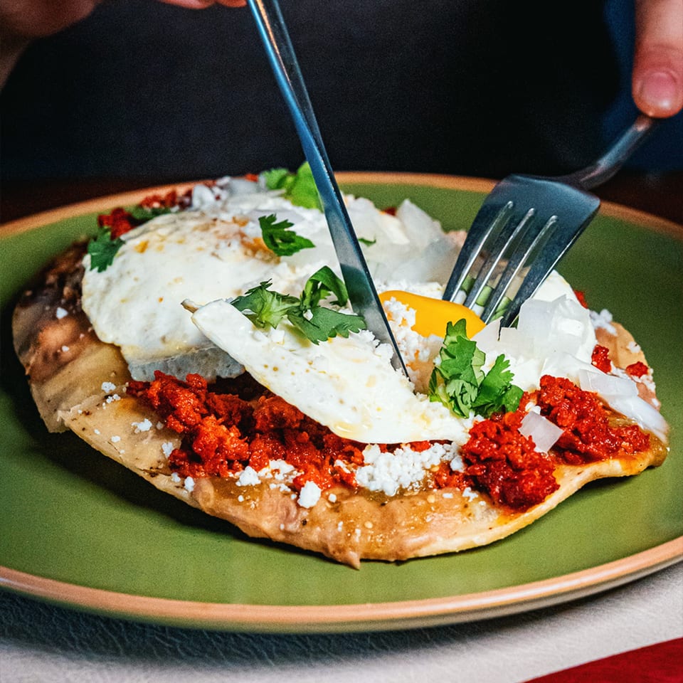 A person cuts into a Mexican breakfast dish with eggs, chorizo, and cheese on a green plate.